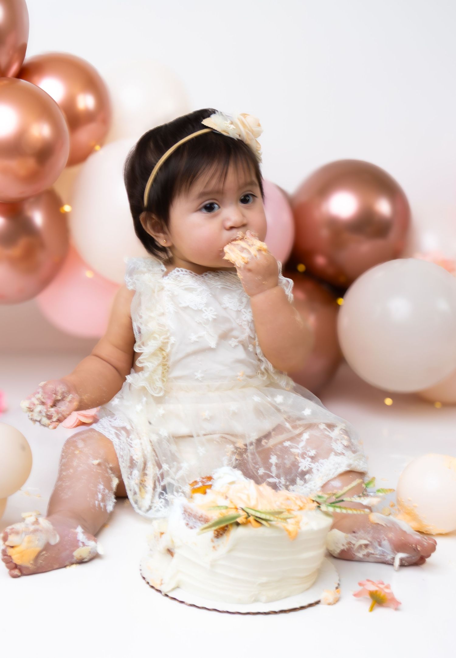 Adorable baby girl in white dress covered in cake during first birthday cake smash photo session.