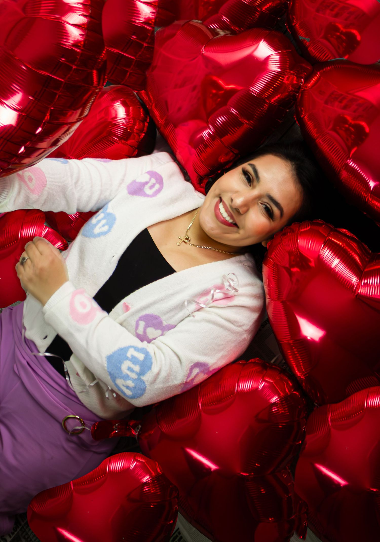 Smiling young woman lying among red heart-shaped foil balloons on the floor.