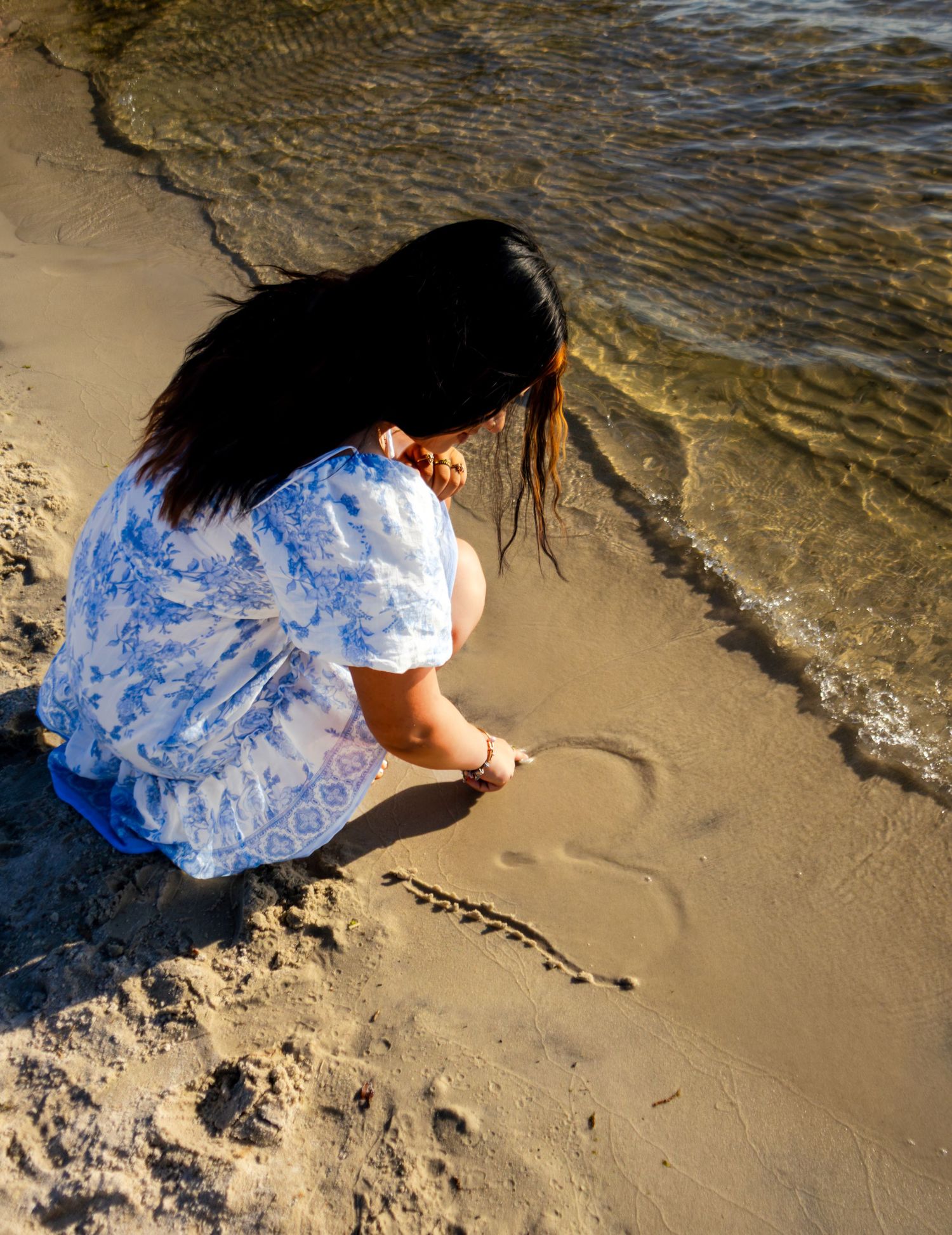 Young woman in blue floral dress writing heart in sand at beach shoreline during golden hour.