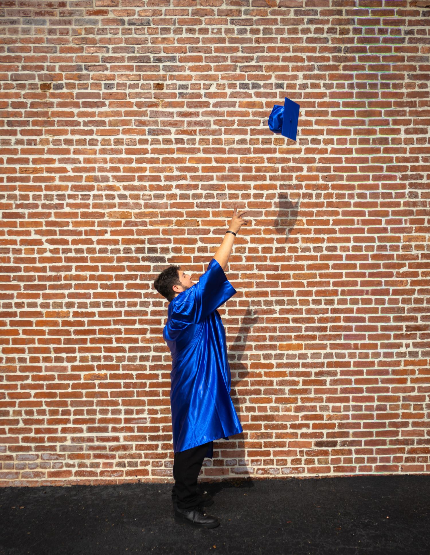 Graduate in blue cap and gown tossing mortarboard into the air against a red brick wall.