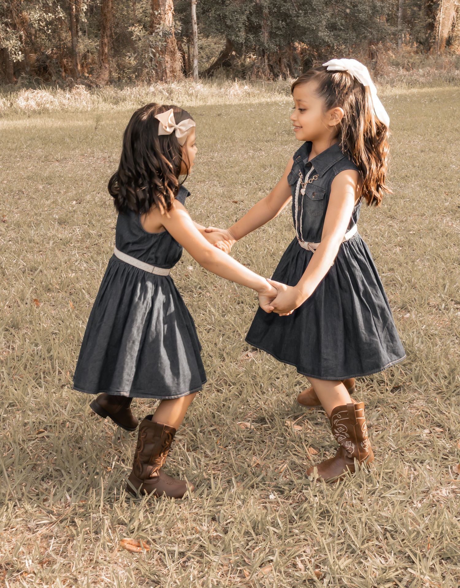 Two young girls in matching denim dresses and cowboy boots hold hands and spin together in a grassy field.