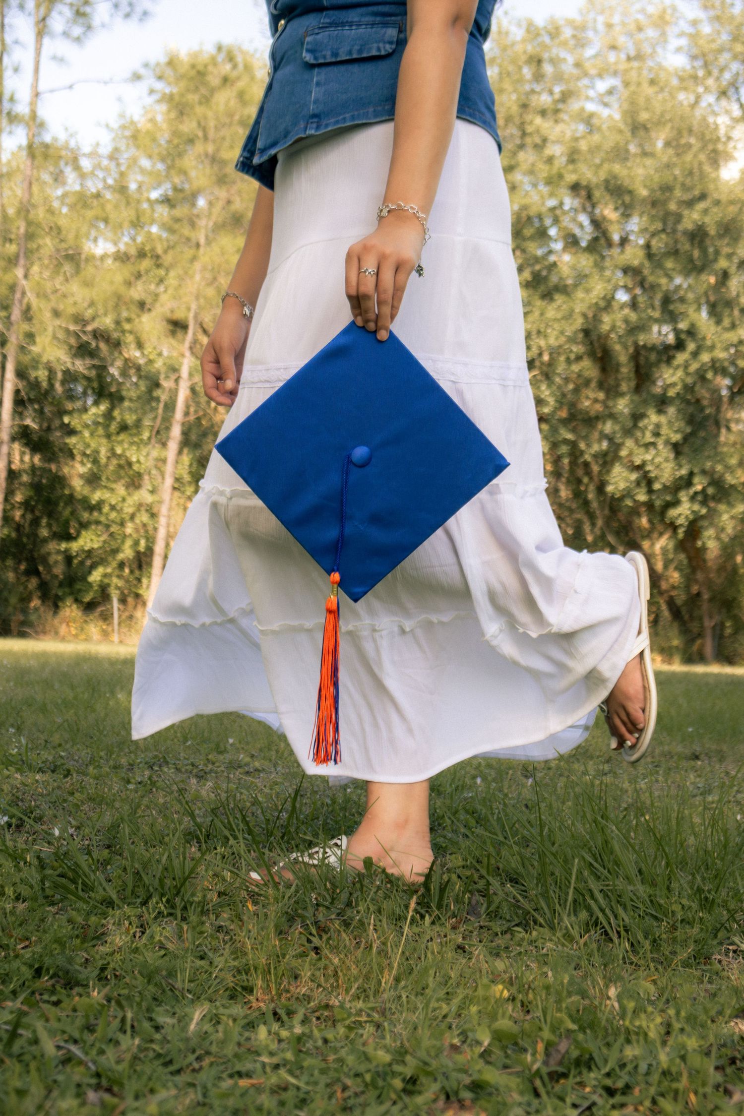Barefoot graduate holding blue mortarboard cap with orange tassel, wearing white skirt and denim vest outdoors.