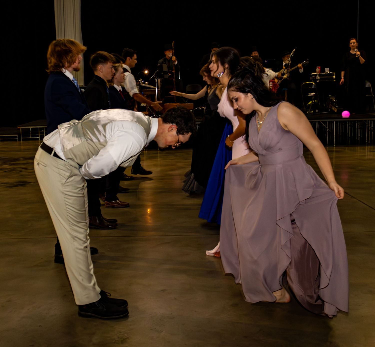 Couple bows to each other during formal dance performance on stage with orchestra behind them.