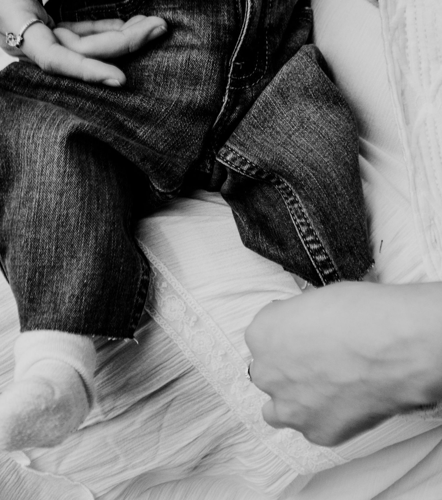 Black and white photo of baby in jeans sitting beside adult in white dress.