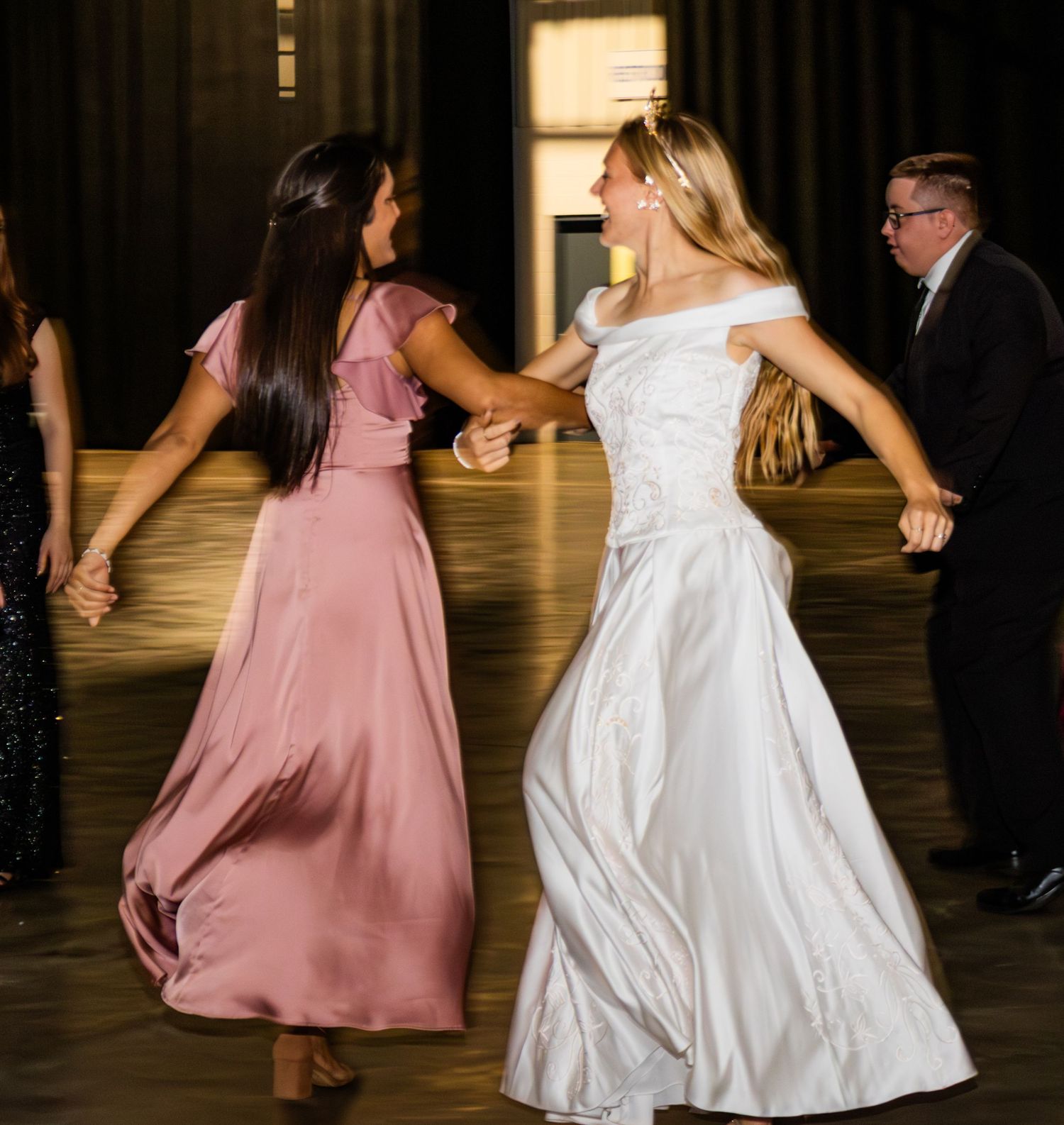Bride in white gown dances joyfully with bridesmaid in pink dress at elegant wedding reception.