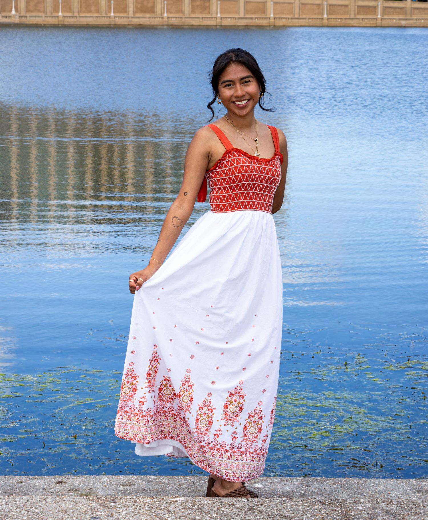 Happy young woman in red and white maxi dress standing by a blue reflective pool.