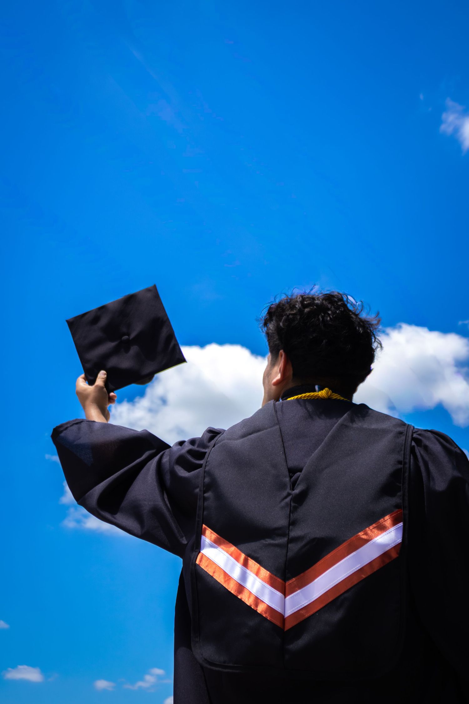 Graduate in black robe holds cap up toward bright blue sky with white clouds, viewed from behind.