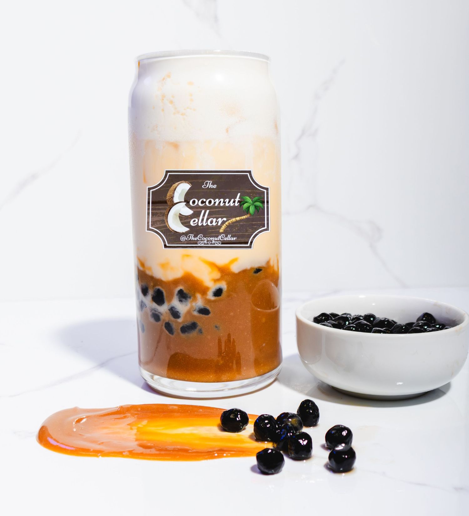 The Coconut Cellar layered boba tea in glass can with tapioca pearls and caramel sauce on marble surface.