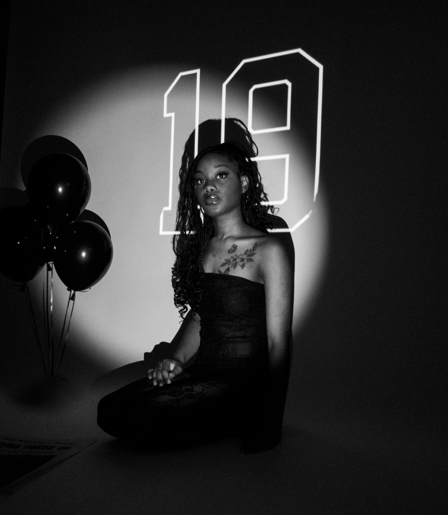 Black and white birthday portrait of a young woman sitting beside balloons with number 19 backdrop.