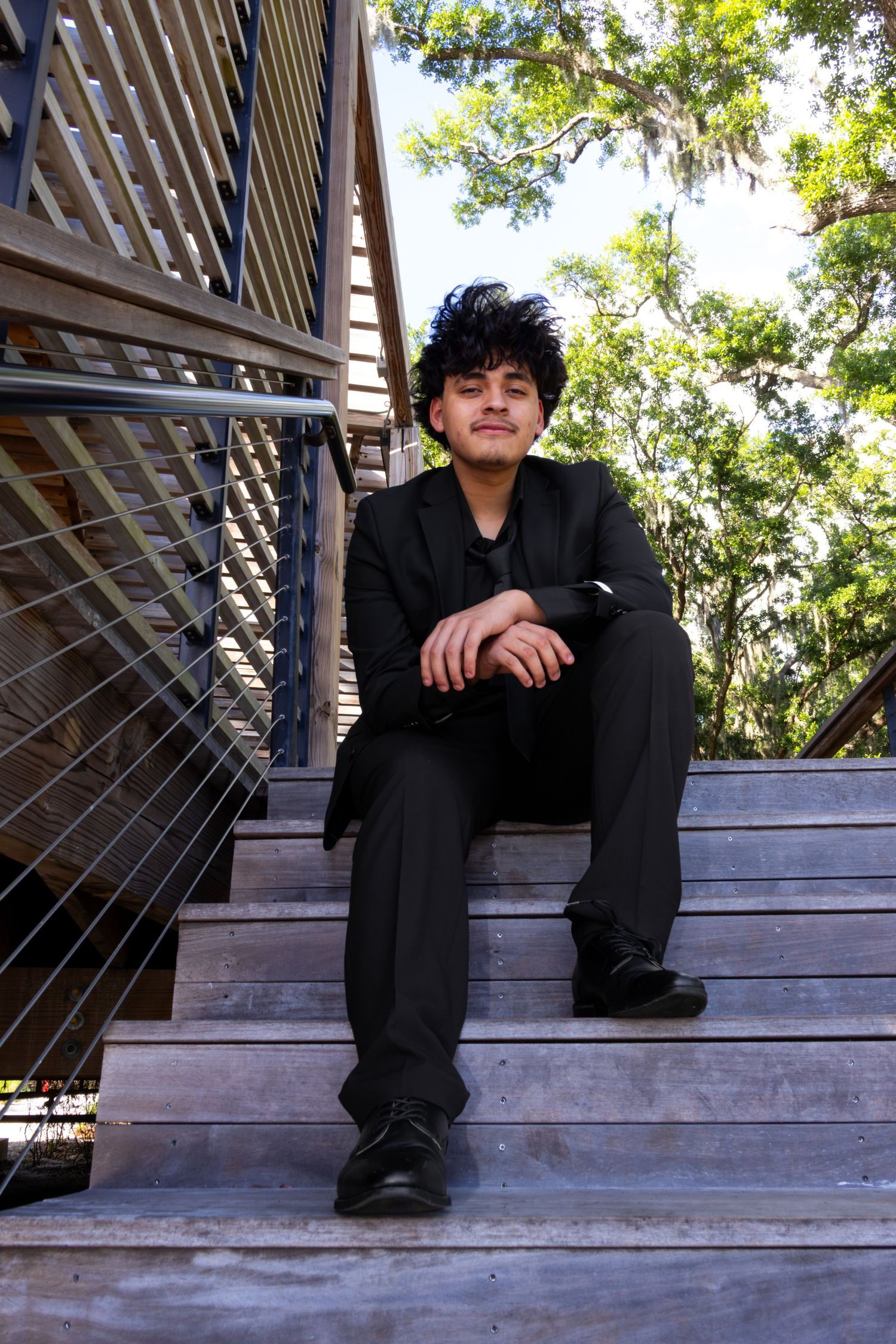 Teenage boy in black suit sits relaxed on wooden outdoor stairs surrounded by lush green trees.