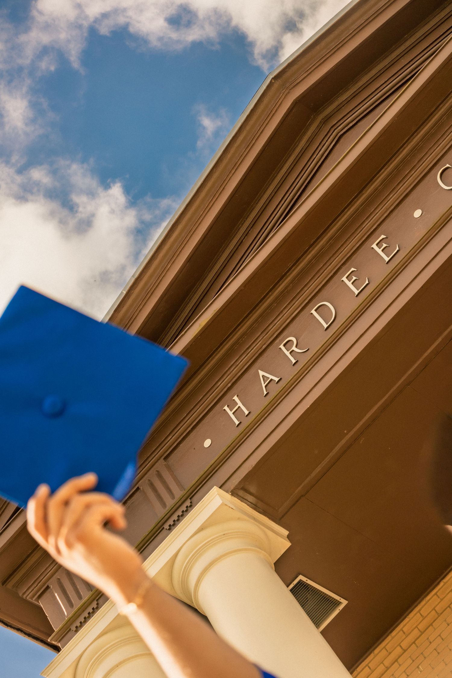 A graduate raises a blue mortarboard cap outside the Hardee building under a partly cloudy sky.