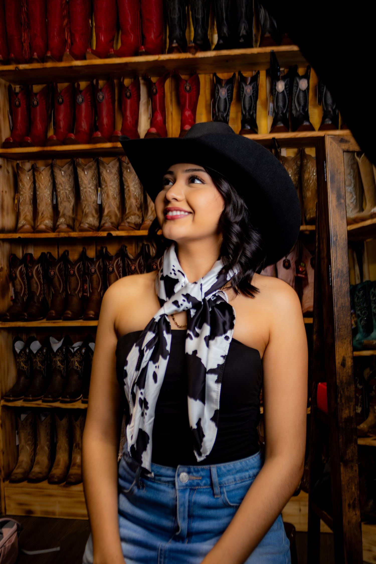 Woman in black cowboy hat and cow print scarf poses in front of wooden shelves displaying colorful cowboy boots.