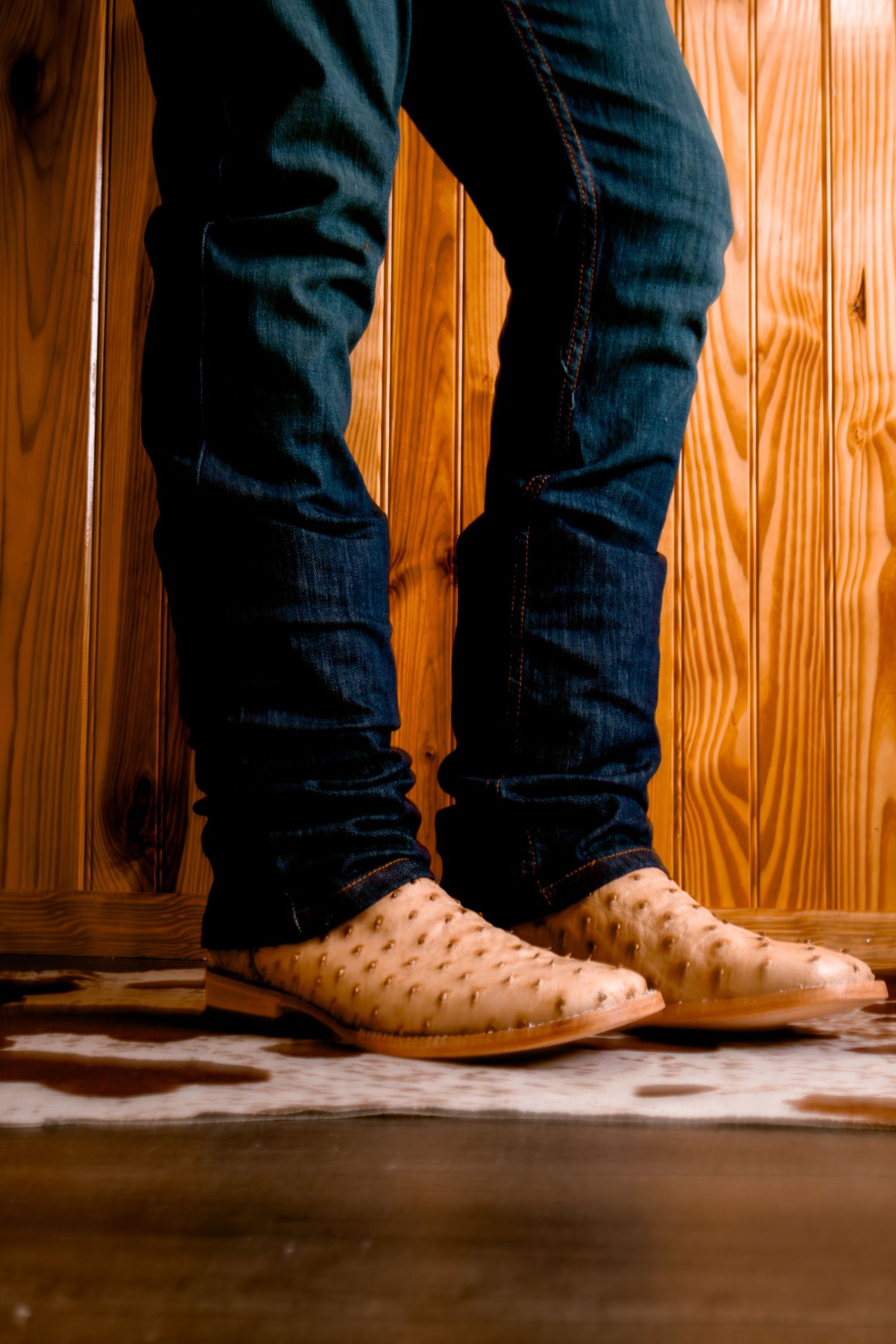 Person wearing tan ostrich leather cowboy boots with dark denim jeans on cowhide rug.