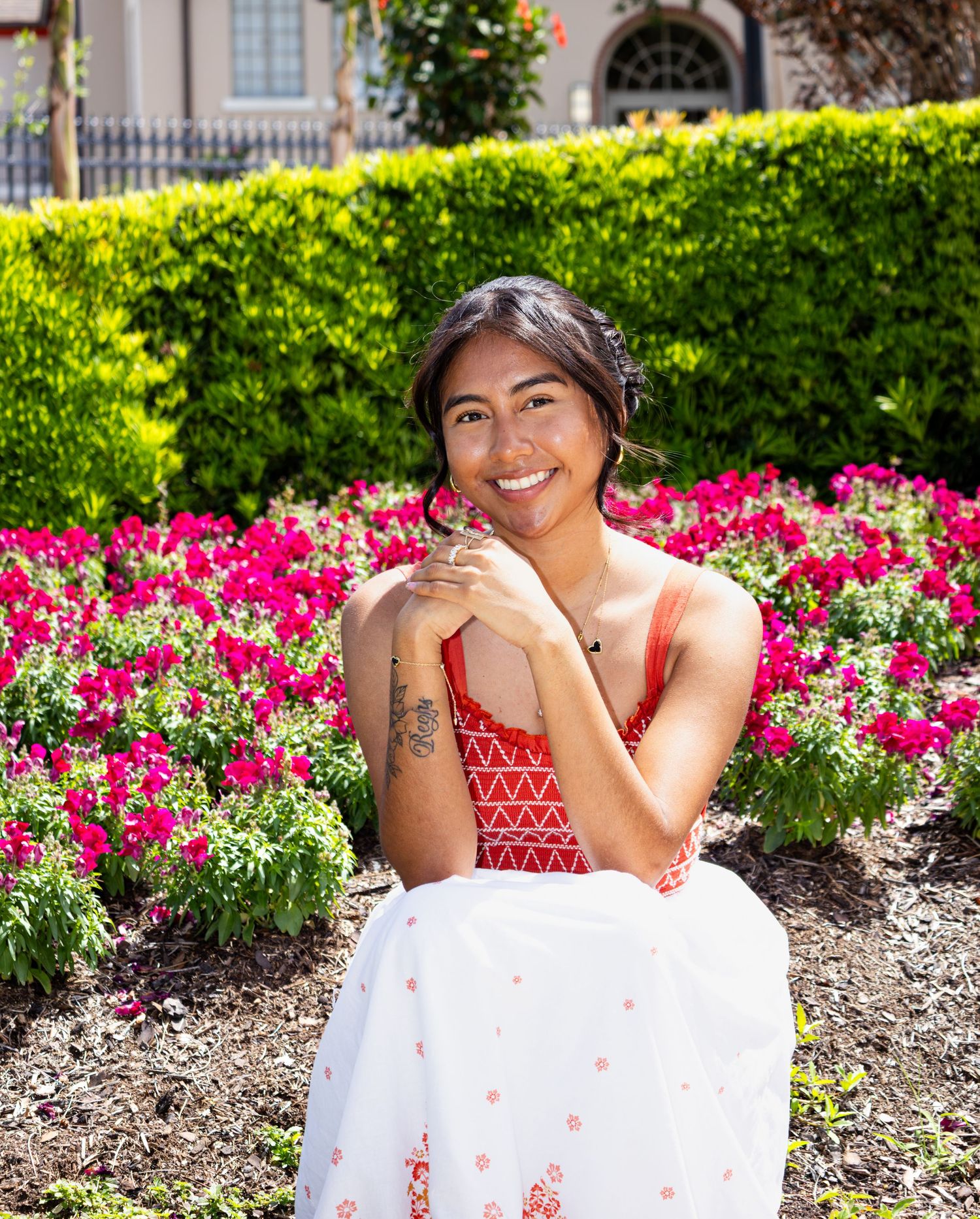 Smiling young woman in red and white dress posing in front of vibrant flower garden.
