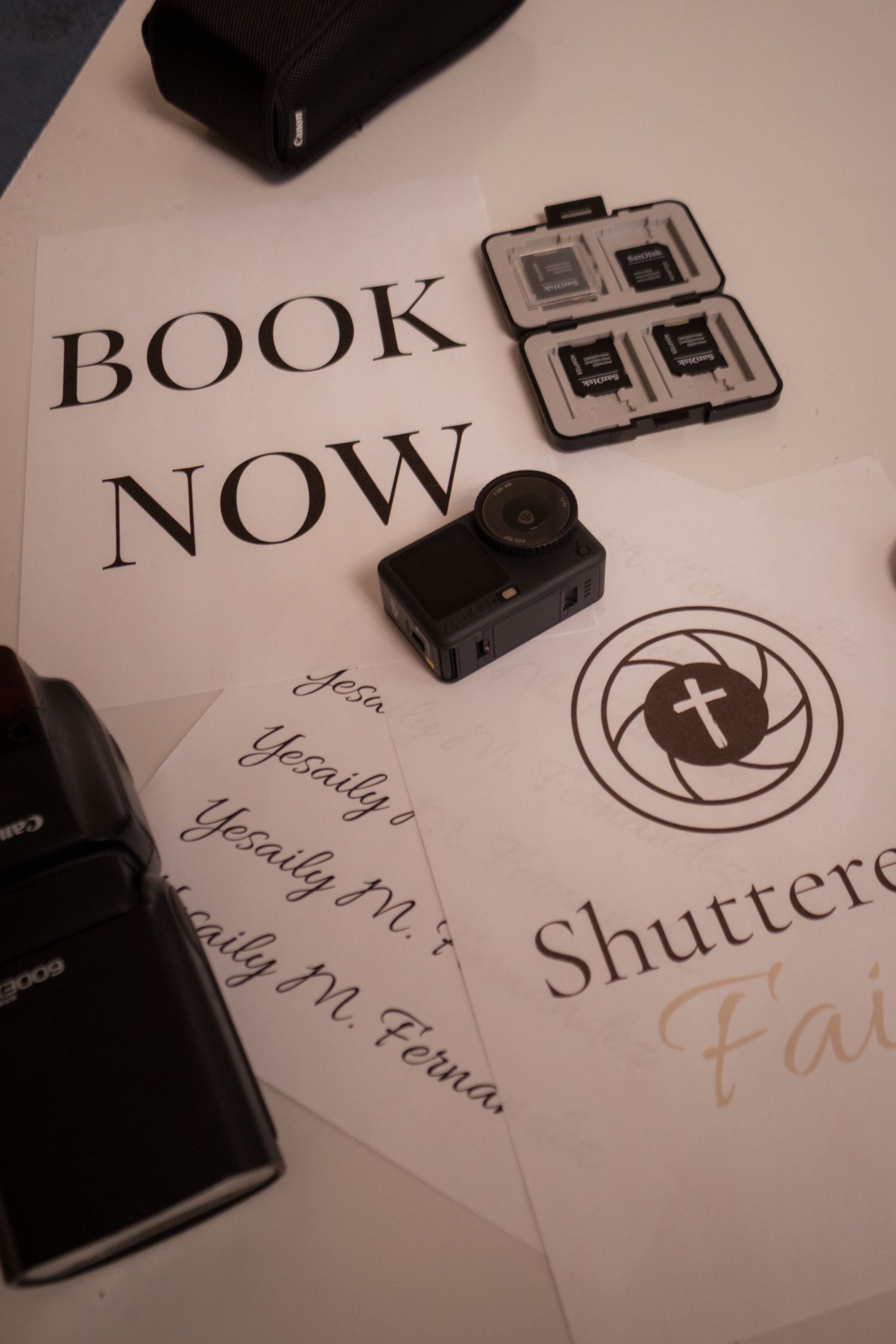 Photography booking display with Canon flash, memory card case, action cam, and Shuttered Faith branded materials on white surface.