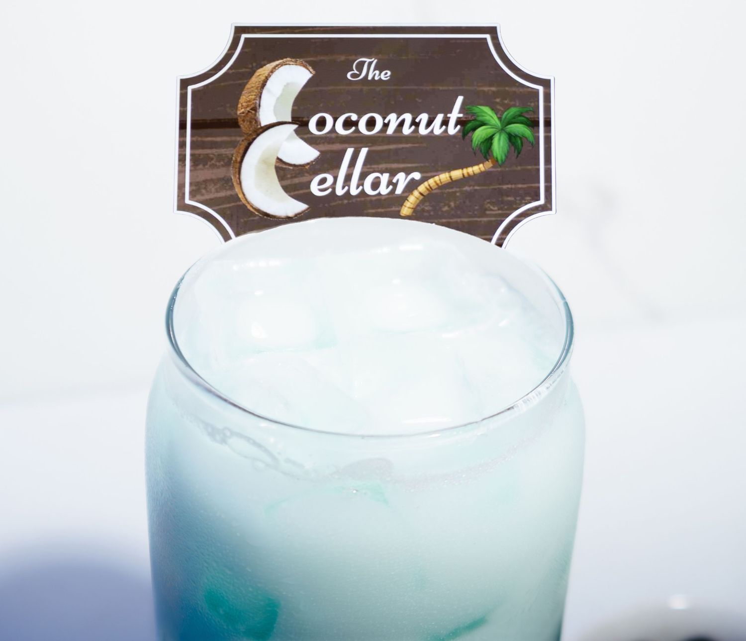 The Coconut Cellar tropical drink in a tall glass with ice and blue hue, garnished with branded wooden sign.