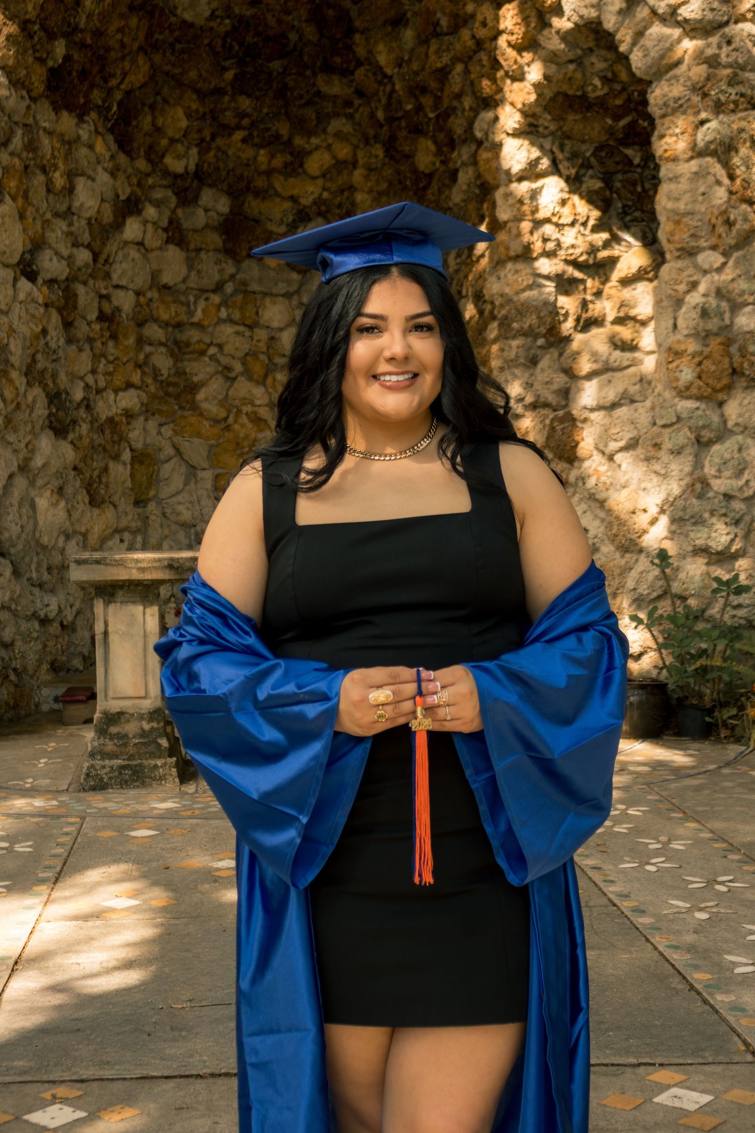 Smiling graduate in blue cap and gown over black dress poses outdoors near stone wall holding red tassel.