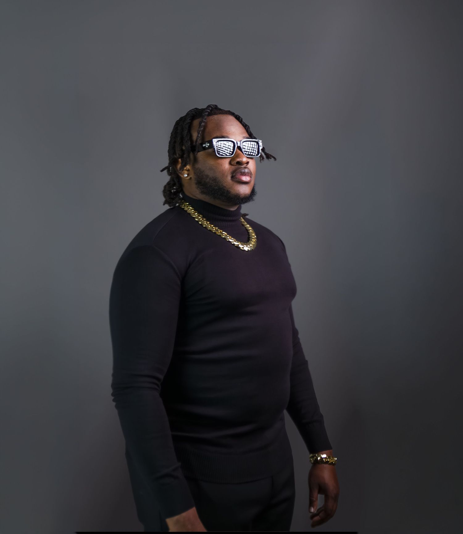 Stylish man in black turtleneck with gold chain necklace and white sunglasses posing against gray background.