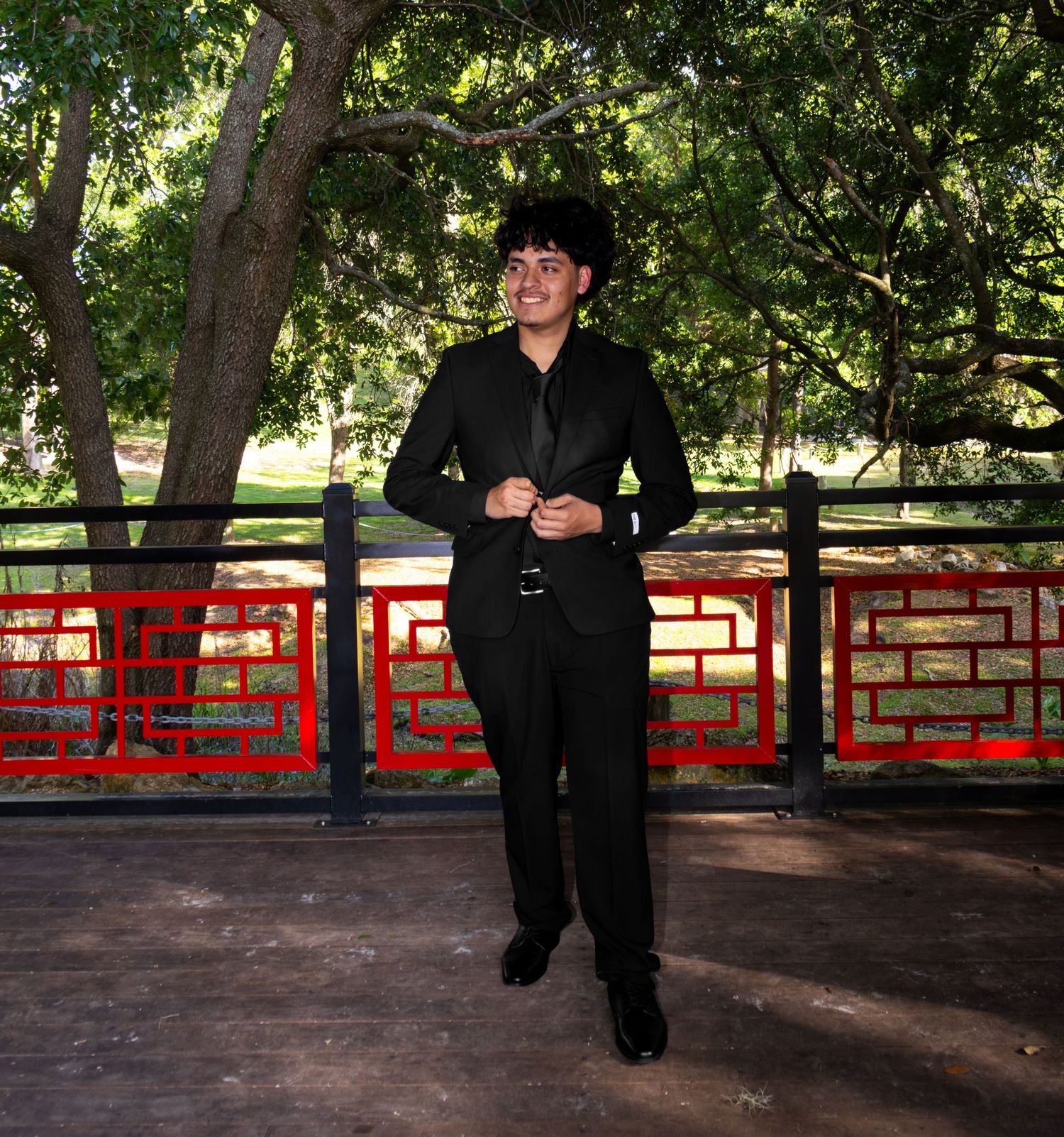 Smiling young man in black suit posing on wooden bridge with red railing in park.