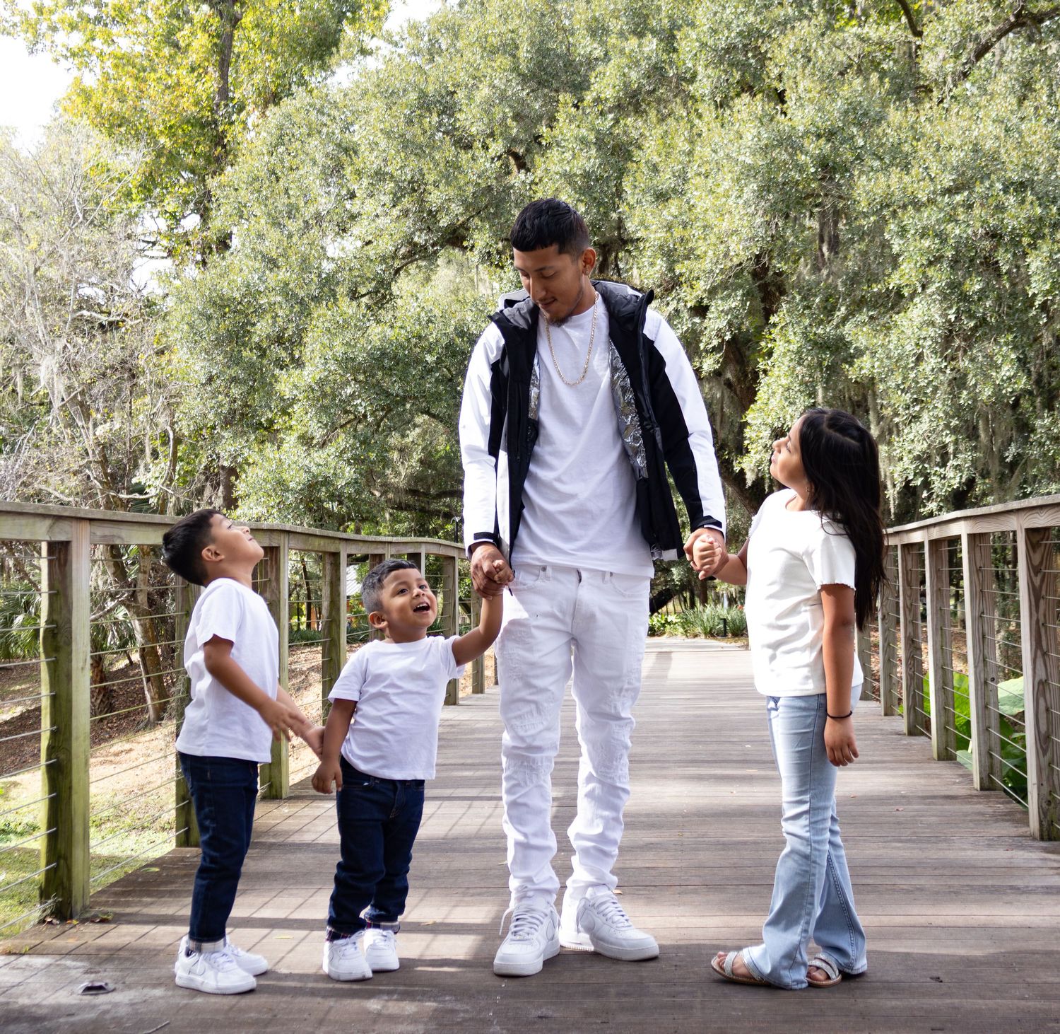Father in white outfit holding hands with three young children on a wooden bridge surrounded by trees.