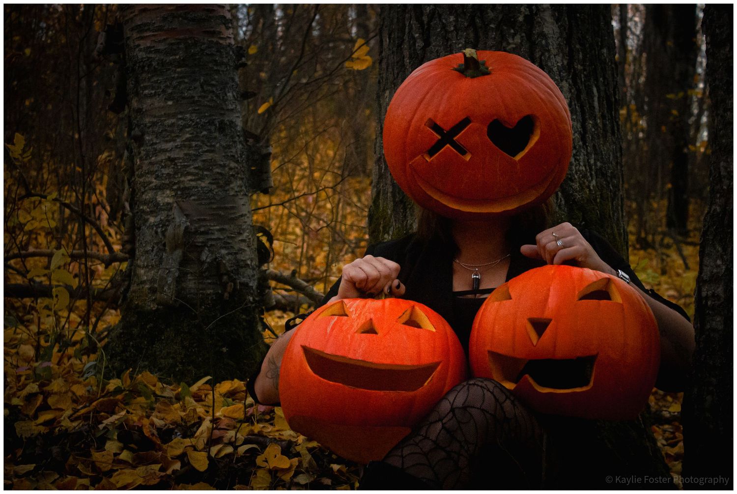 Person holding Halloween jack-o-lanterns in autumn forest, with a pumpkin covering their face.
