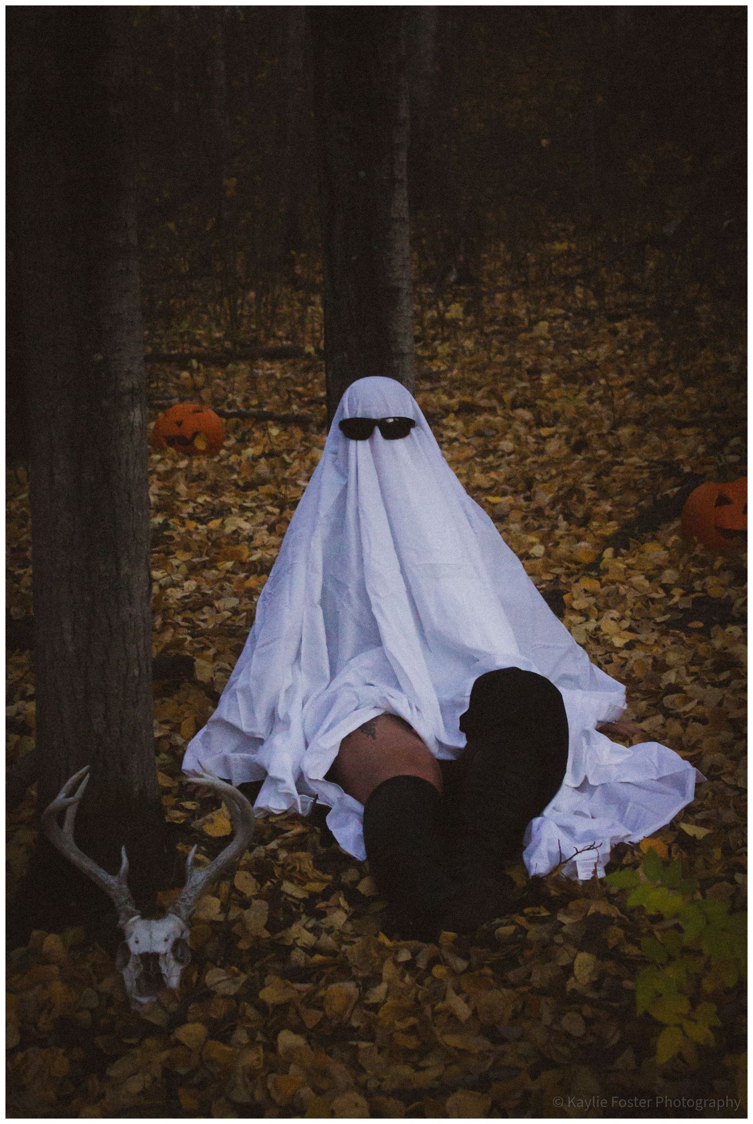 Person in white ghost costume with sunglasses sitting among autumn leaves in forest with pumpkins and deer skull.