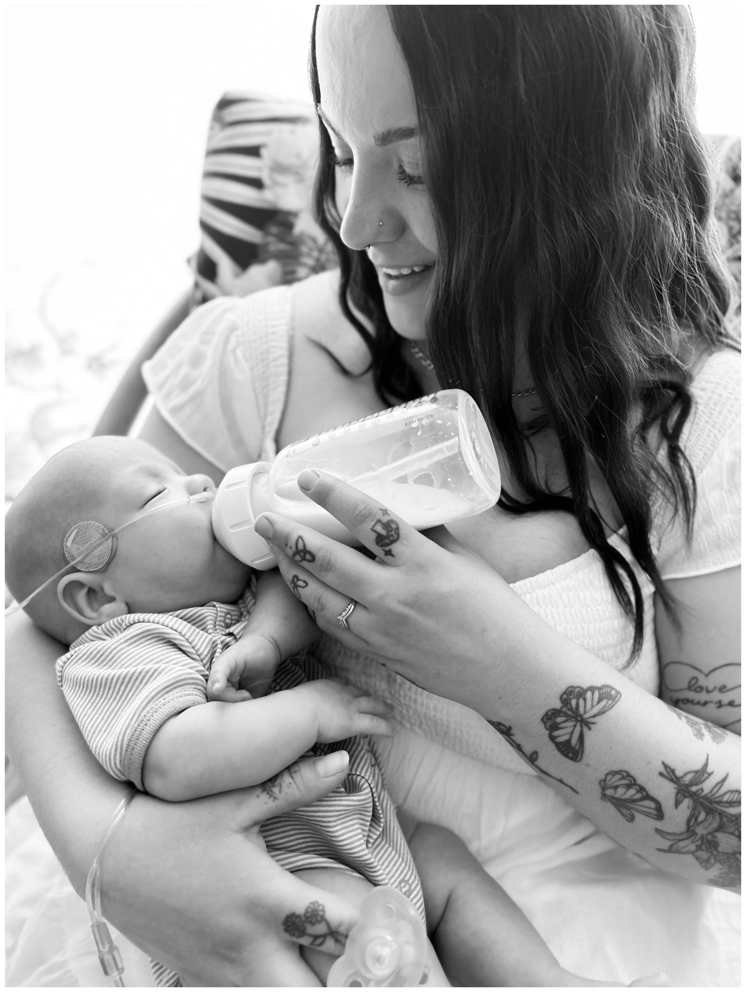 Black and white photo of a tattooed mother smiling while bottle feeding her newborn baby.