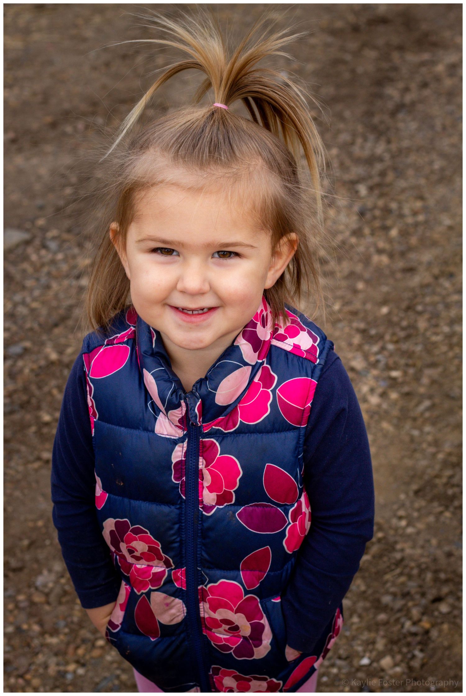 Adorable smiling toddler girl with ponytail wearing navy floral puffer vest outdoors.