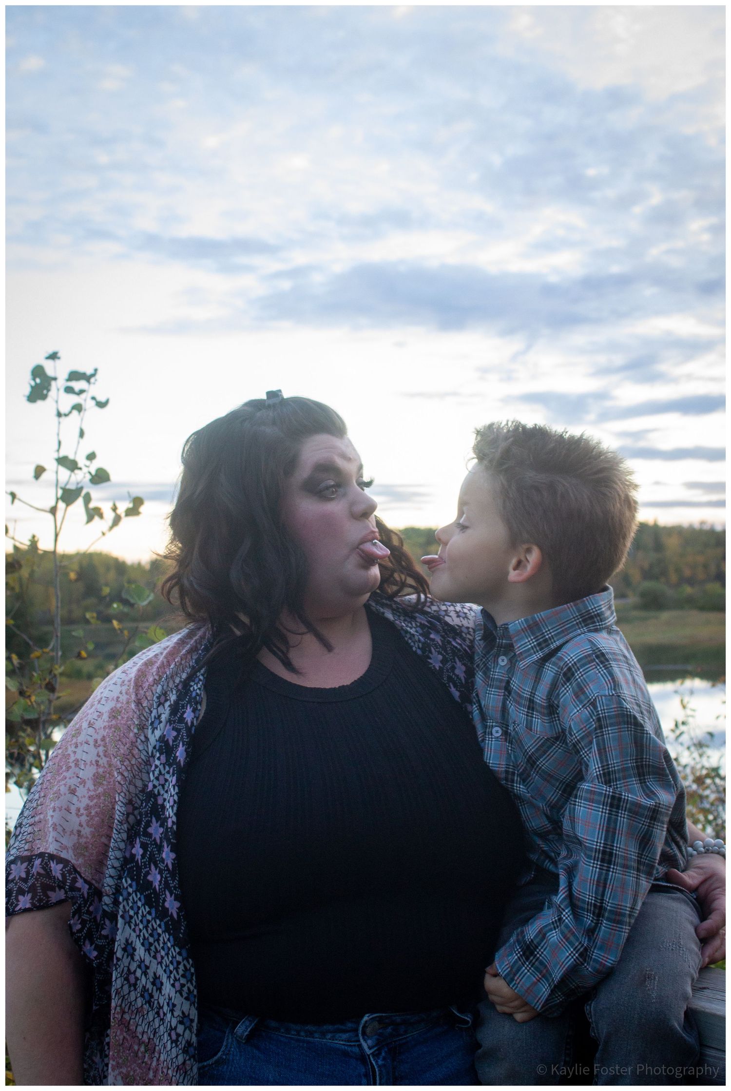 A woman and young boy share a tender moment outdoors near a lake at sunset.
