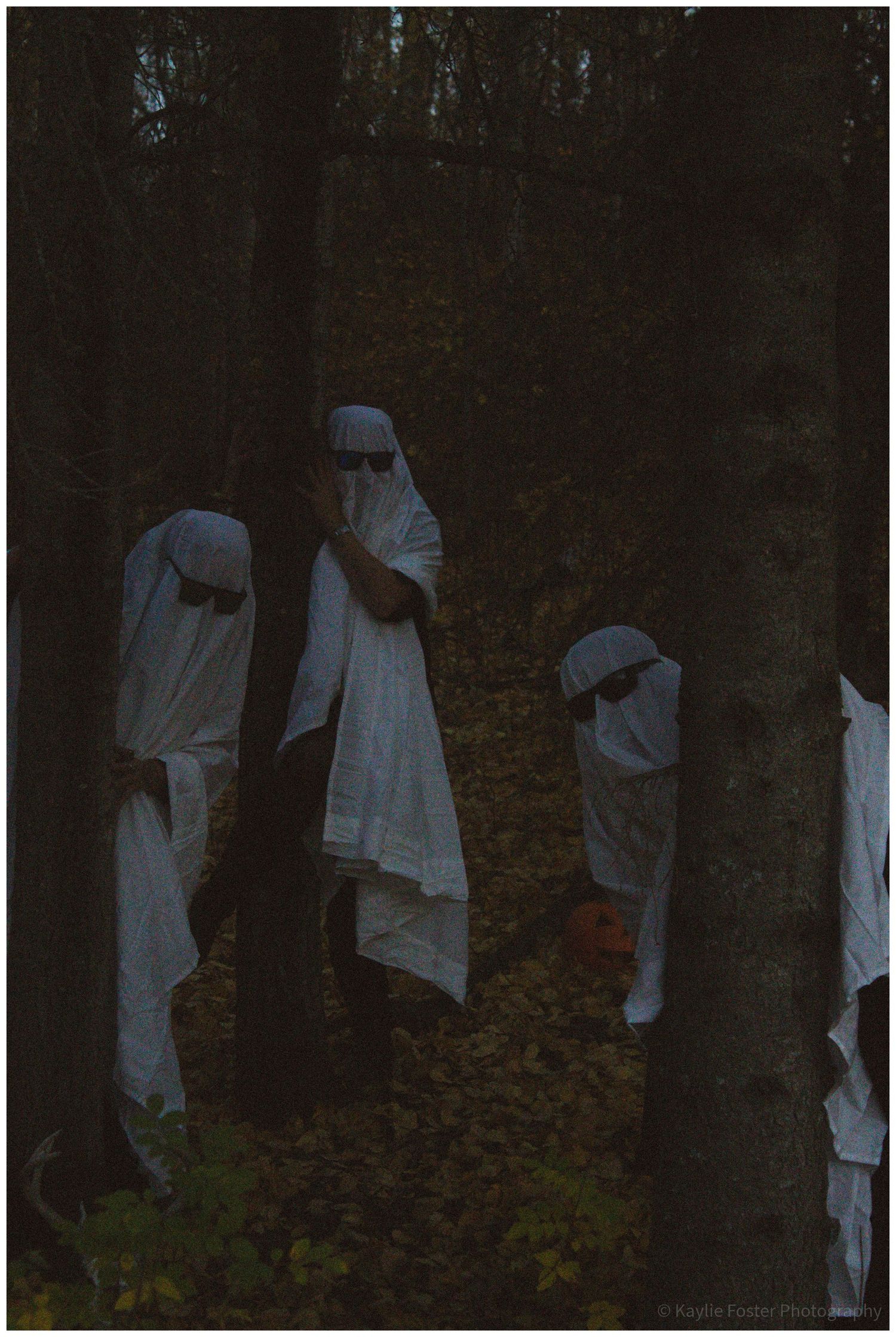 Children dressed as ghosts in white sheets standing among trees in a dark forest on Halloween.