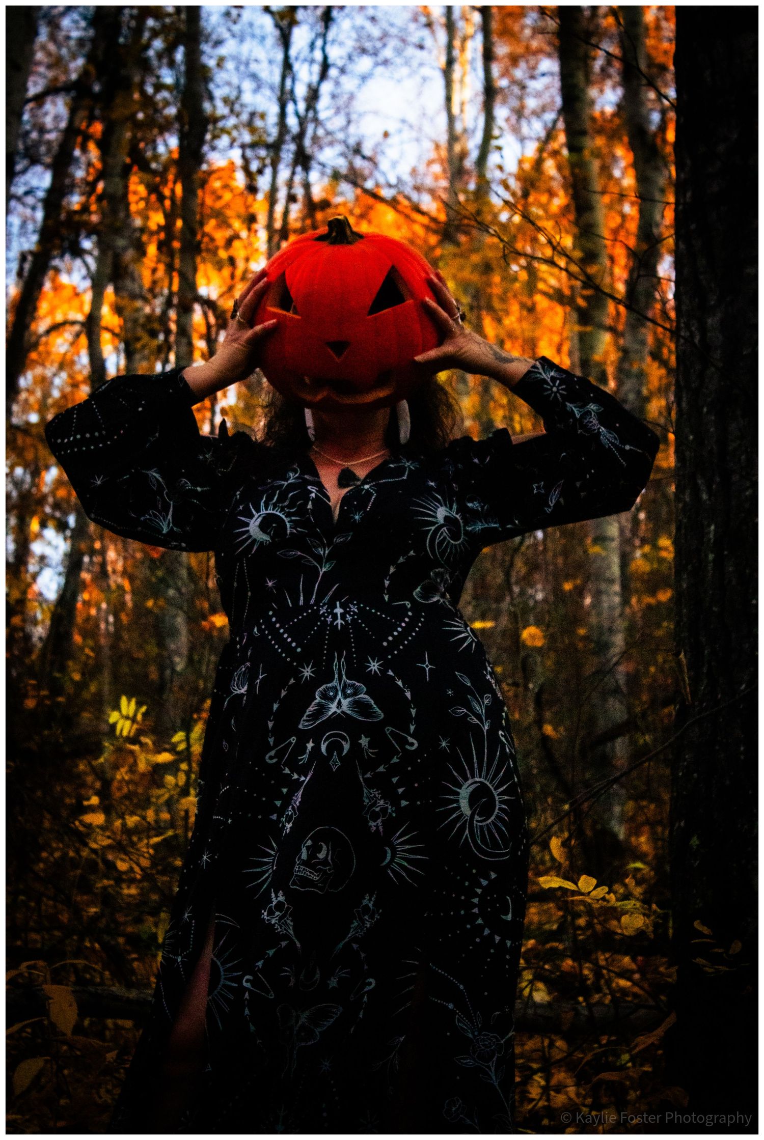 A woman in a black celestial print dress holds a carved jack-o-lantern over her face in an autumn forest.