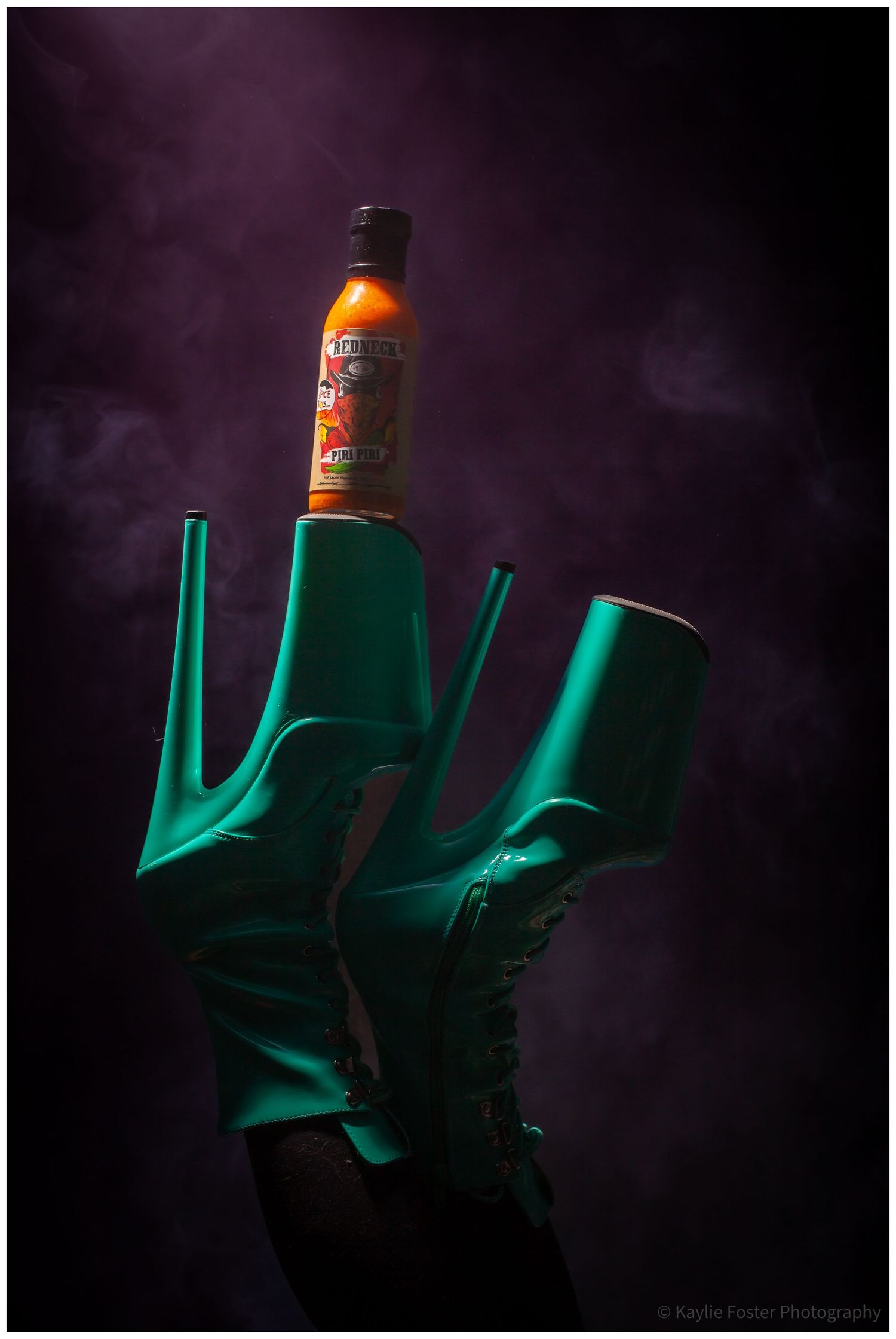 Teal platform heel boots holding an orange hot sauce bottle with purple smoky background.