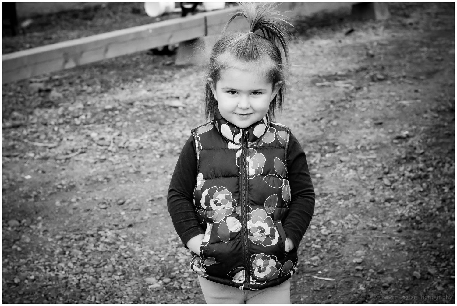 Black and white portrait of young girl with ponytail in floral vest standing on dirt path.