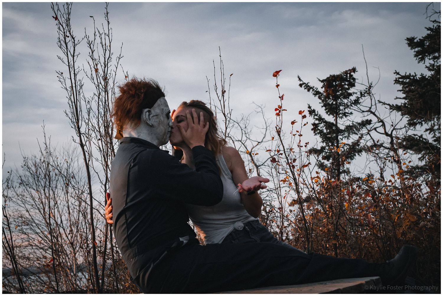 Couple in Halloween costume sitting outdoors, one wearing Michael Myers mask, sharing romantic moment against autumn trees.