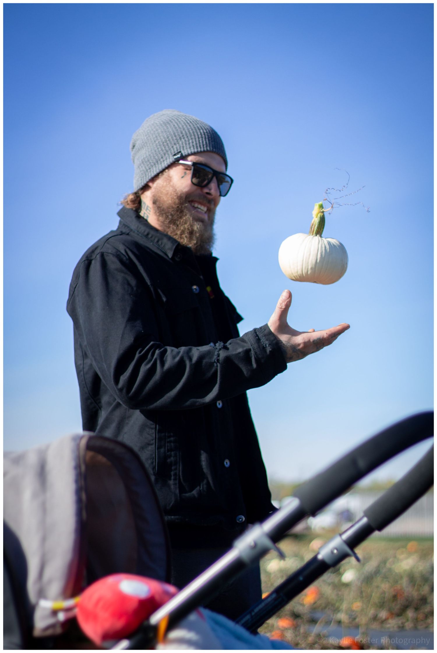 Bearded man in gray beanie tossing a white pumpkin in the air outdoors on a sunny day.