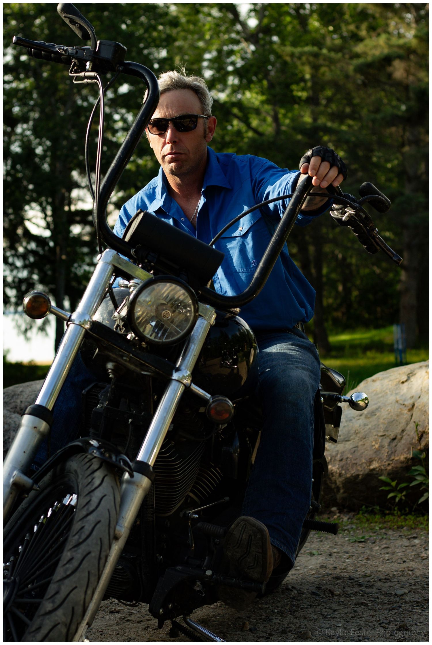 Man in blue shirt and sunglasses sitting on a classic motorcycle outdoors.