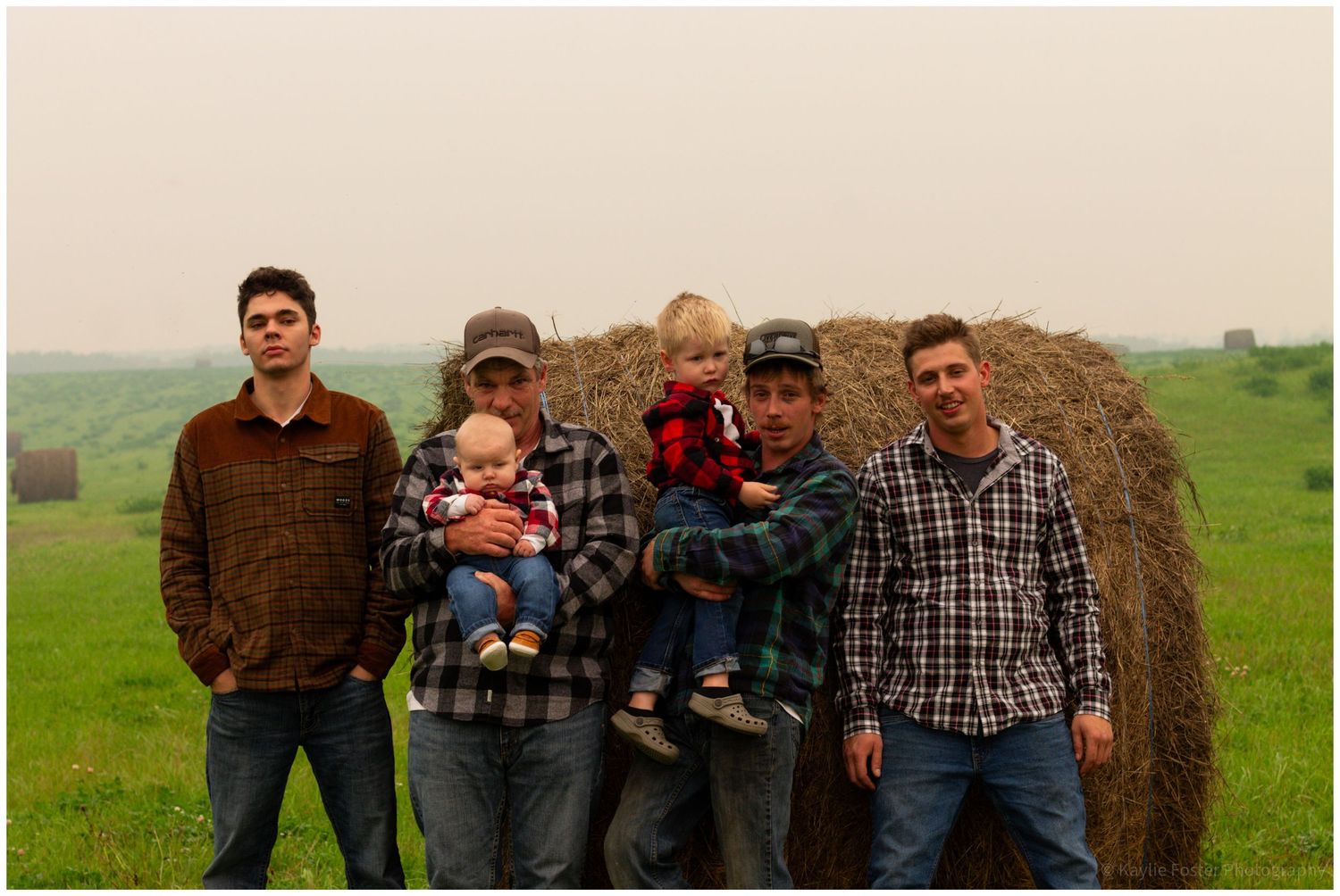 Group of four men and two toddlers pose together in front of a hay bale in a field.