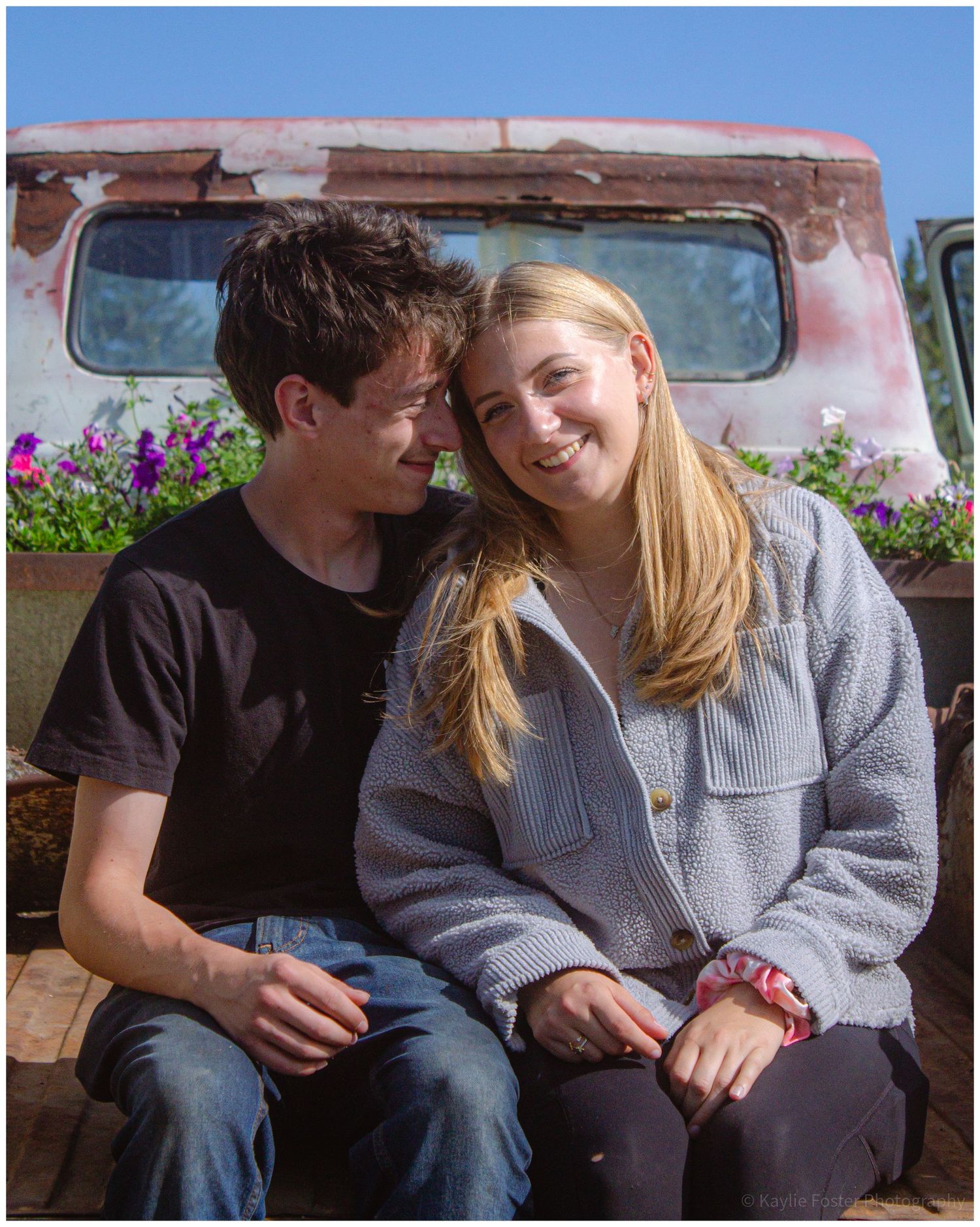 A young couple sits together smiling in front of a rustic vintage truck with purple flowers on a sunny day.