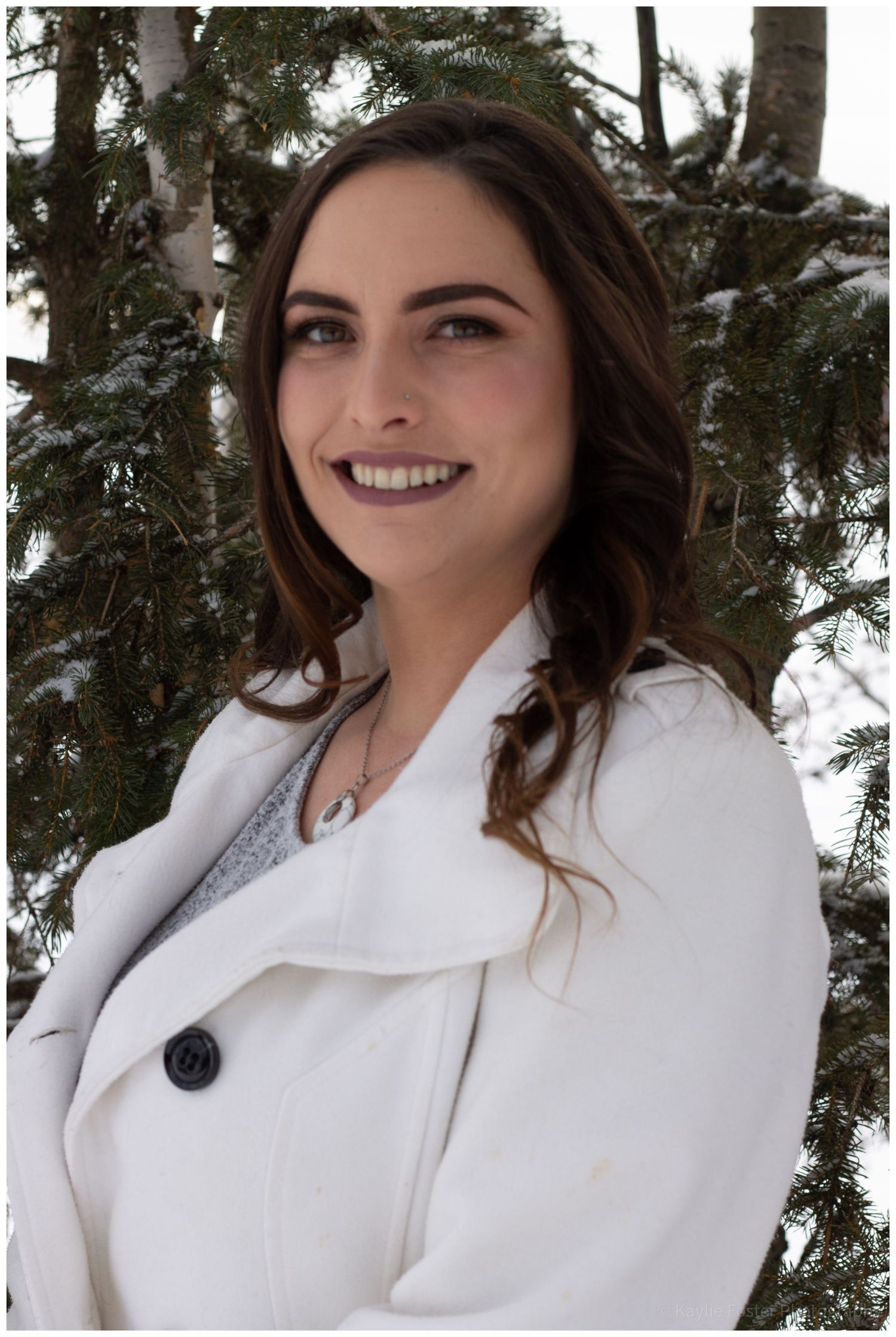 Young woman in white coat smiling outdoors in winter, snow-covered evergreen trees in background.