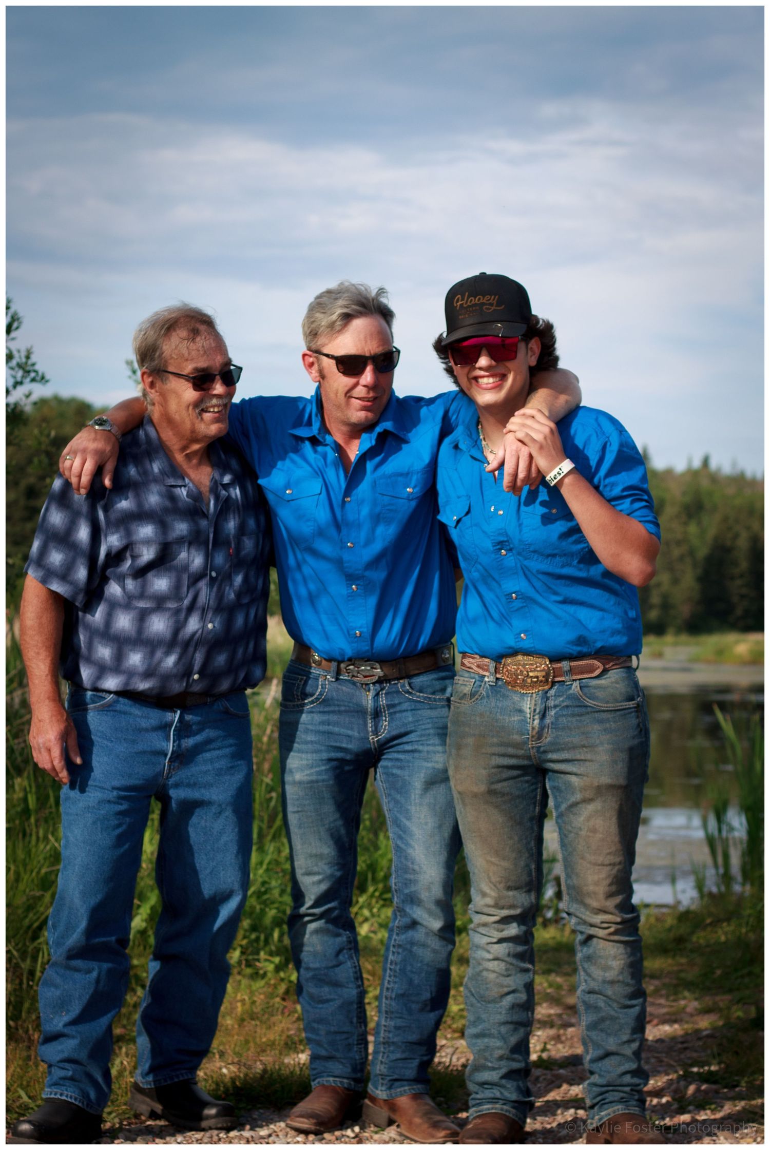Three men in blue shirts smiling with arms around each other near a pond.
