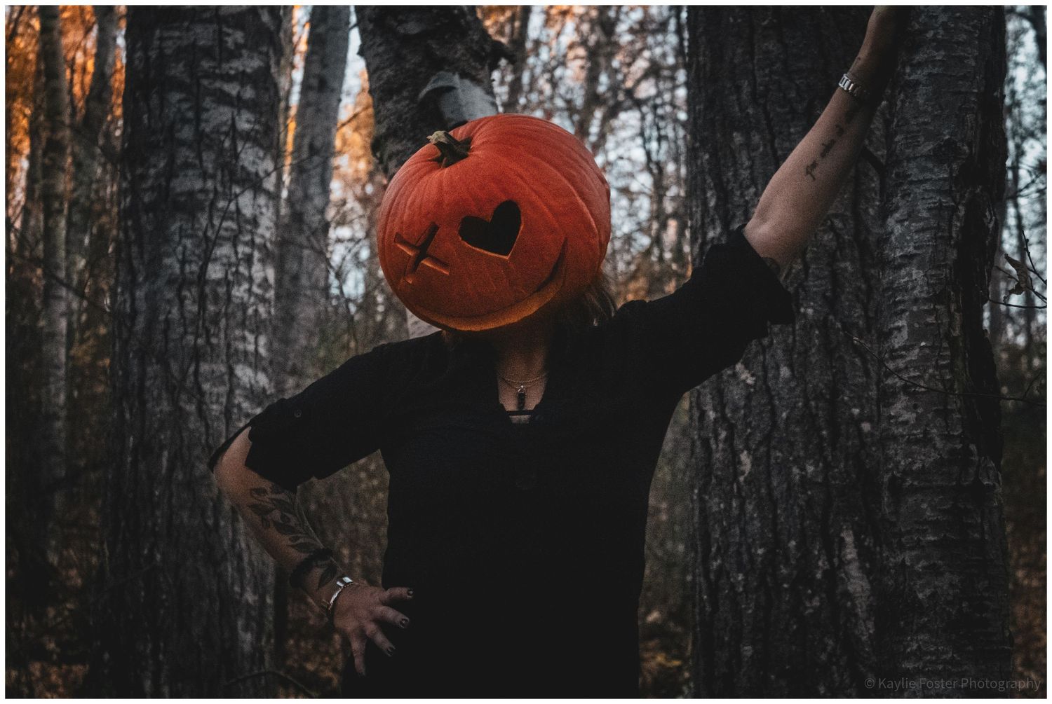 A person in black clothing poses with a carved Halloween pumpkin over their head in a dark autumn forest.