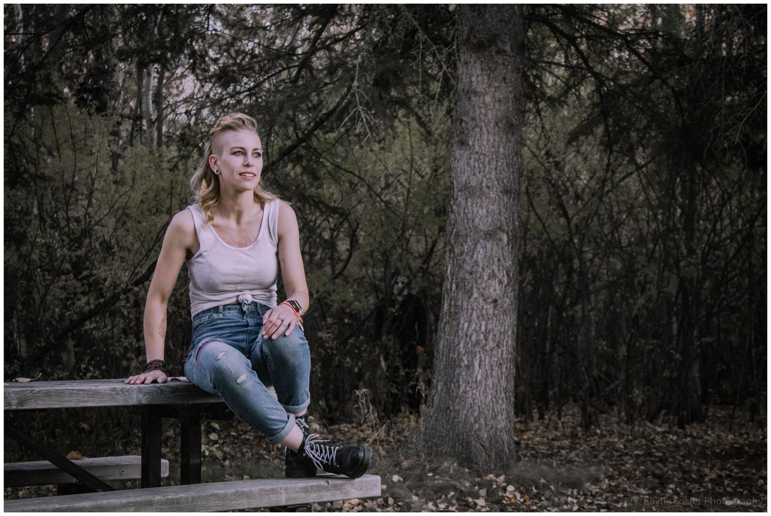 Young woman in jeans and tank top sits on wooden picnic table in a moody forest setting.
