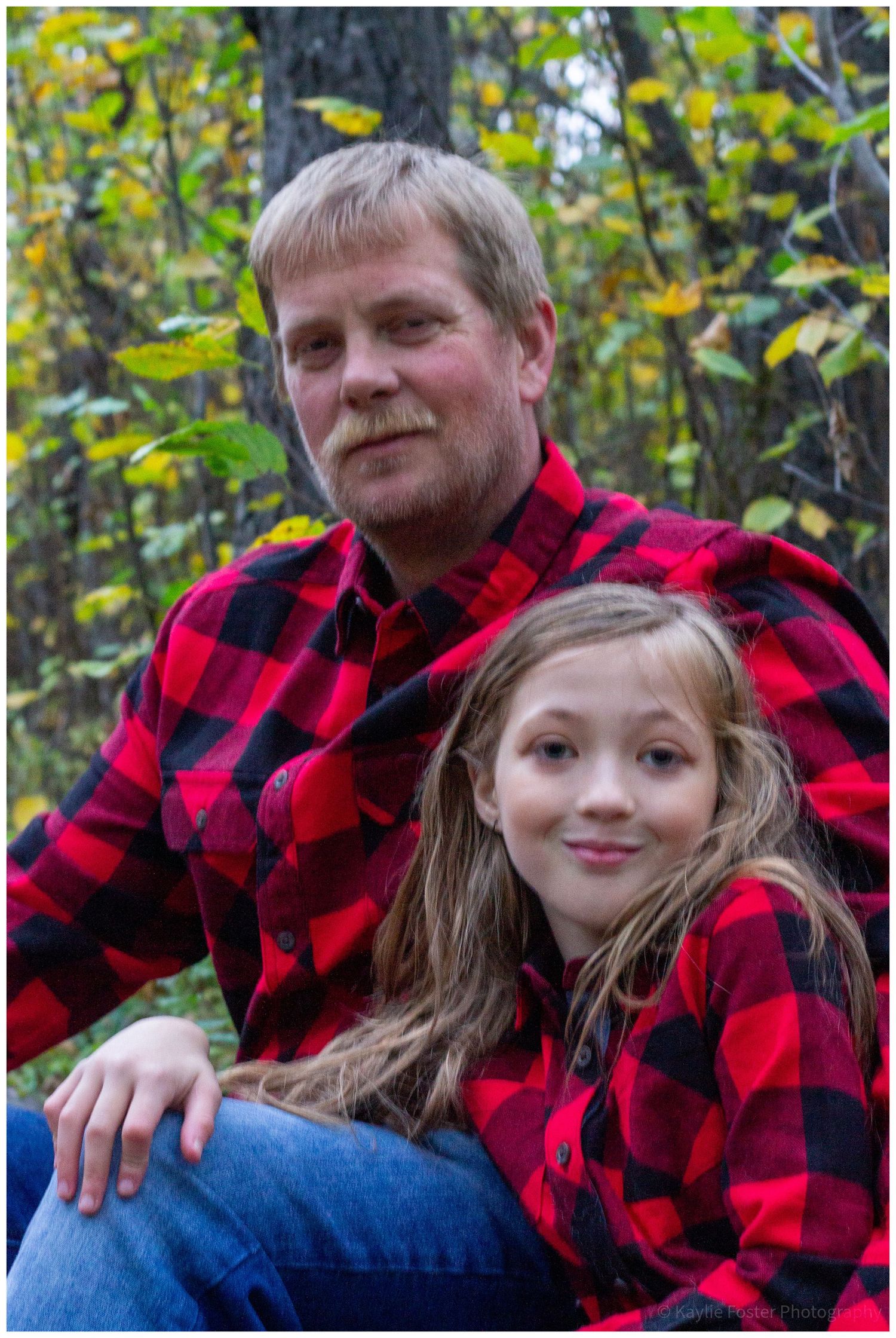 Father and daughter in matching red plaid shirts sitting together in autumn forest.
