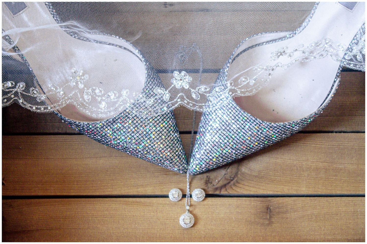 Sparkling crystal bridal heels with pearl drop earrings resting on wooden floor beneath delicate lace wedding veil.