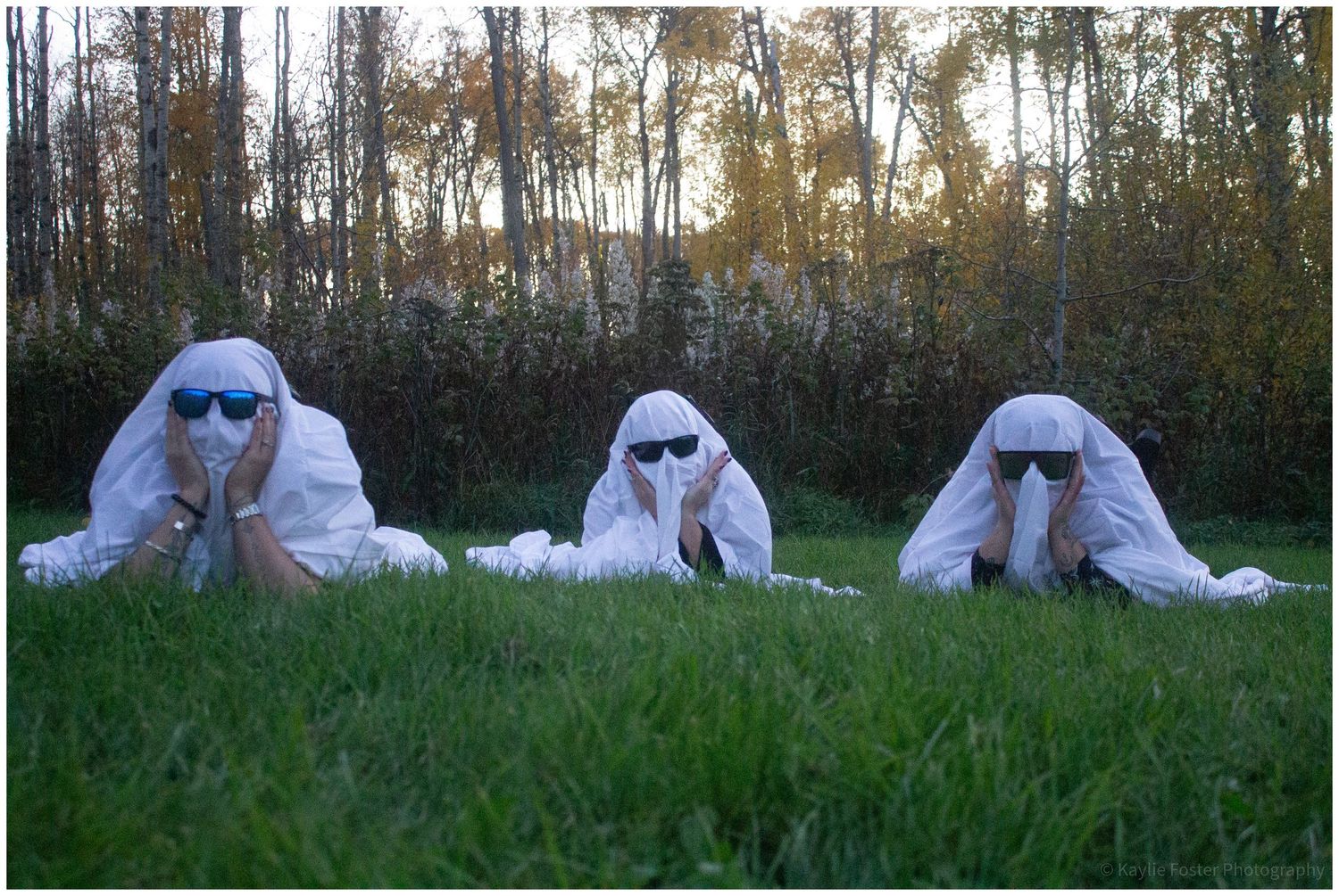 Three people in white ghost costumes with sunglasses lying on green grass in front of trees at sunset.