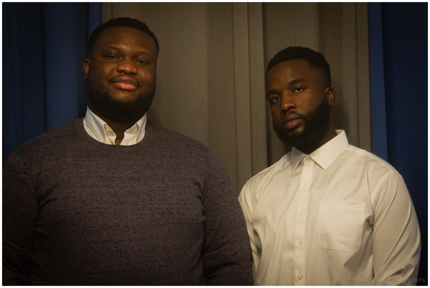 Two men in casual and formal attire posing together indoors against a gray curtain backdrop.