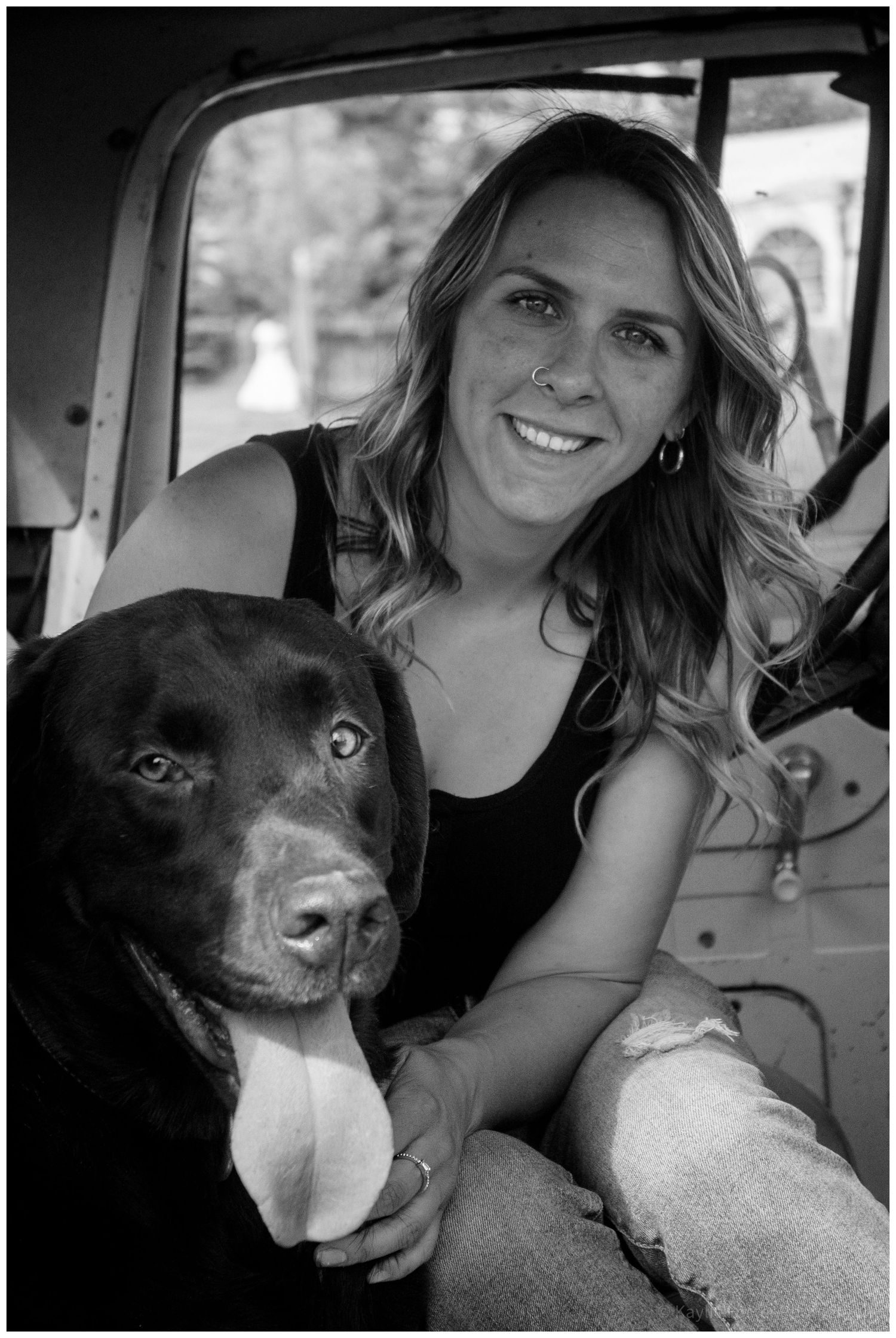 Woman smiling with chocolate Labrador dog inside vintage truck cab, black and white portrait photo.