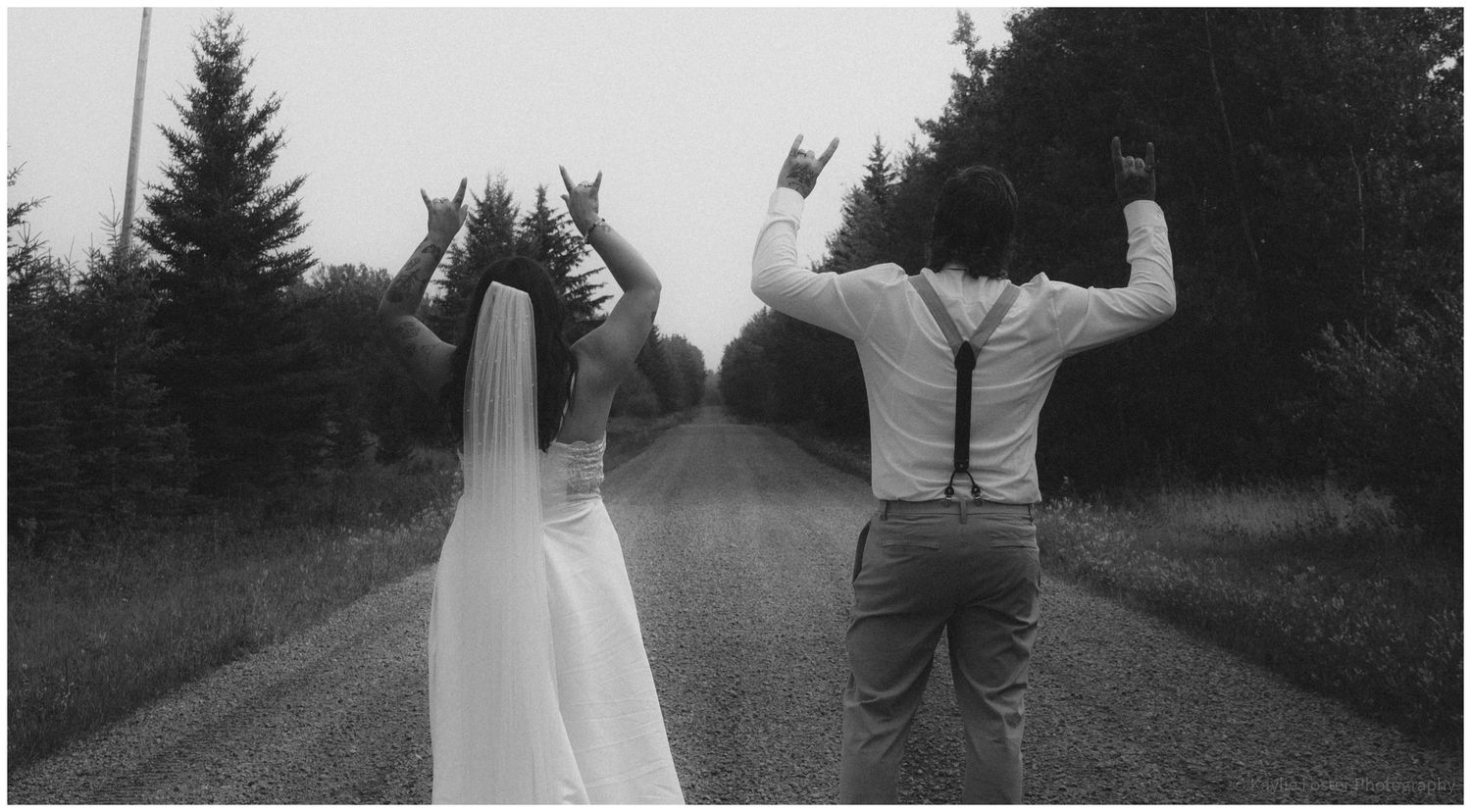 Black and white photo of bride and groom making rock on hand gestures on a tree-lined country road.