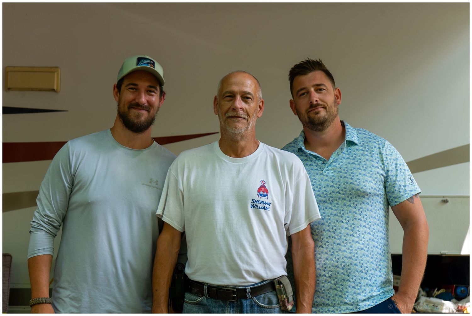 Father and sons posing together indoors, with the middle man wearing a white logo t-shirt.