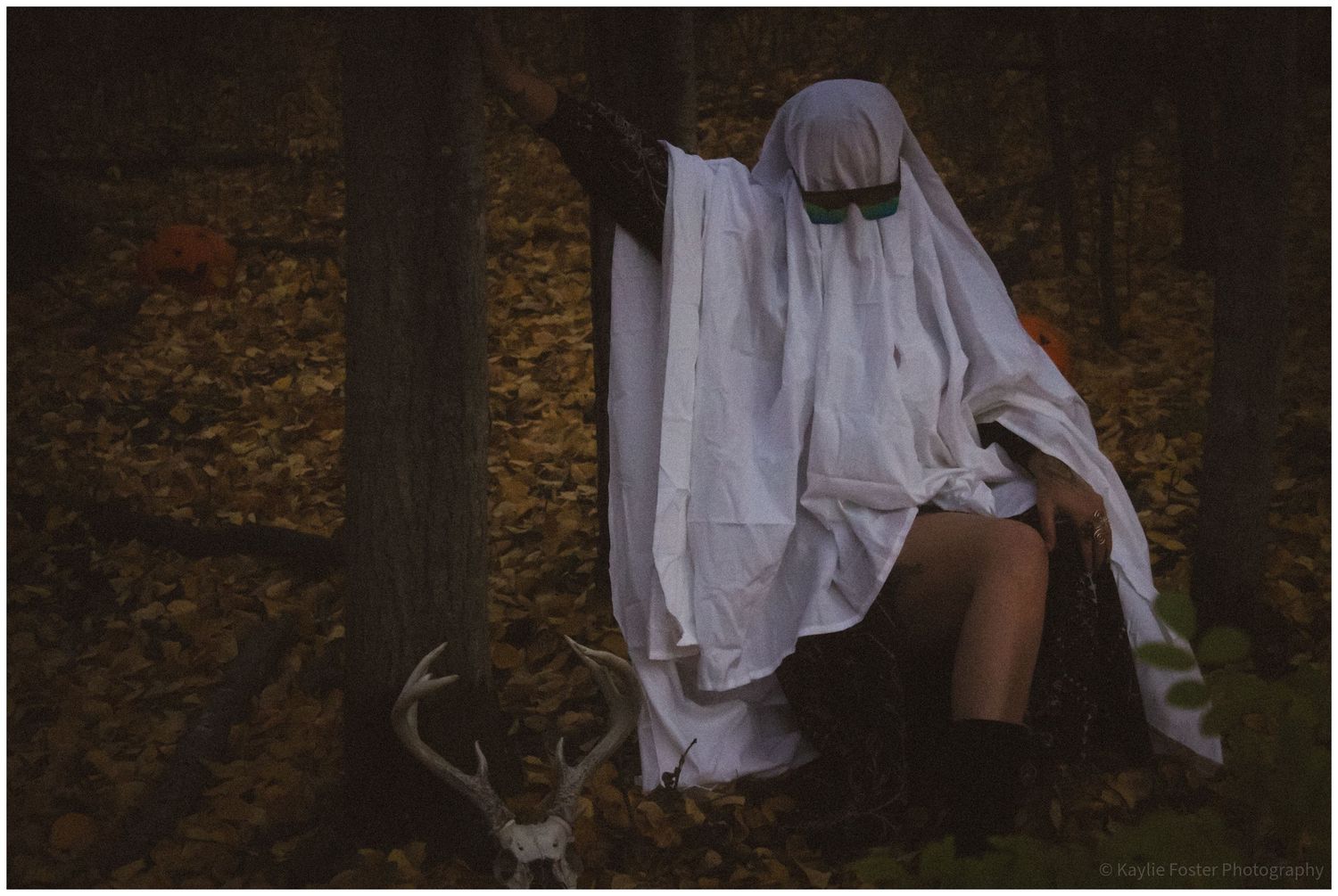 Person dressed as a ghost in white sheet costume sitting in autumn forest with deer antlers nearby.
